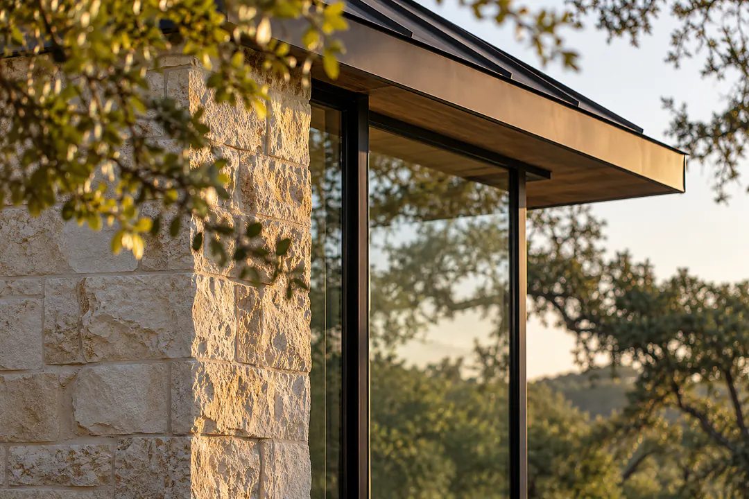 Split-face limestone meeting a black steel window frame under a bronze roof edge, in late sun.